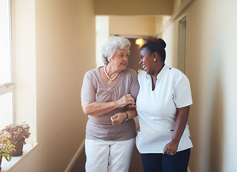 AdobeStock_112047591 Caregiver laughs with elderly woman while escorting her down a hall