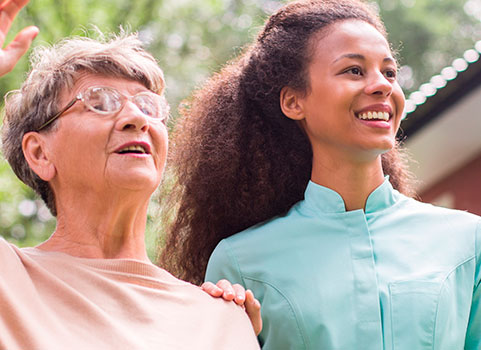 Young female caregiver smiles as elderly woman greets someone in the distance